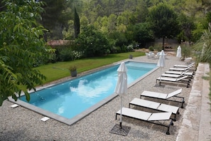 A rectangular swimming pool with a clear blue water surface, surrounded by a pebble border. There are lounge chairs with white umbrellas aligned along the pool's edge.