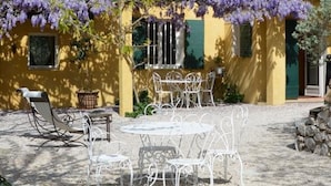 A yellow building with green shutters, a patio with white metal furniture, and purple flowering vines. - Le Mas des Aludes (La Destrousse)