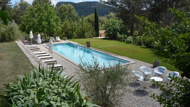 A rectangular swimming pool with a clear blue water, surrounded by a gravel deck. There are lounge chairs and umbrellas along the poolside. - Le Mas des Aludes (La Destrousse)
