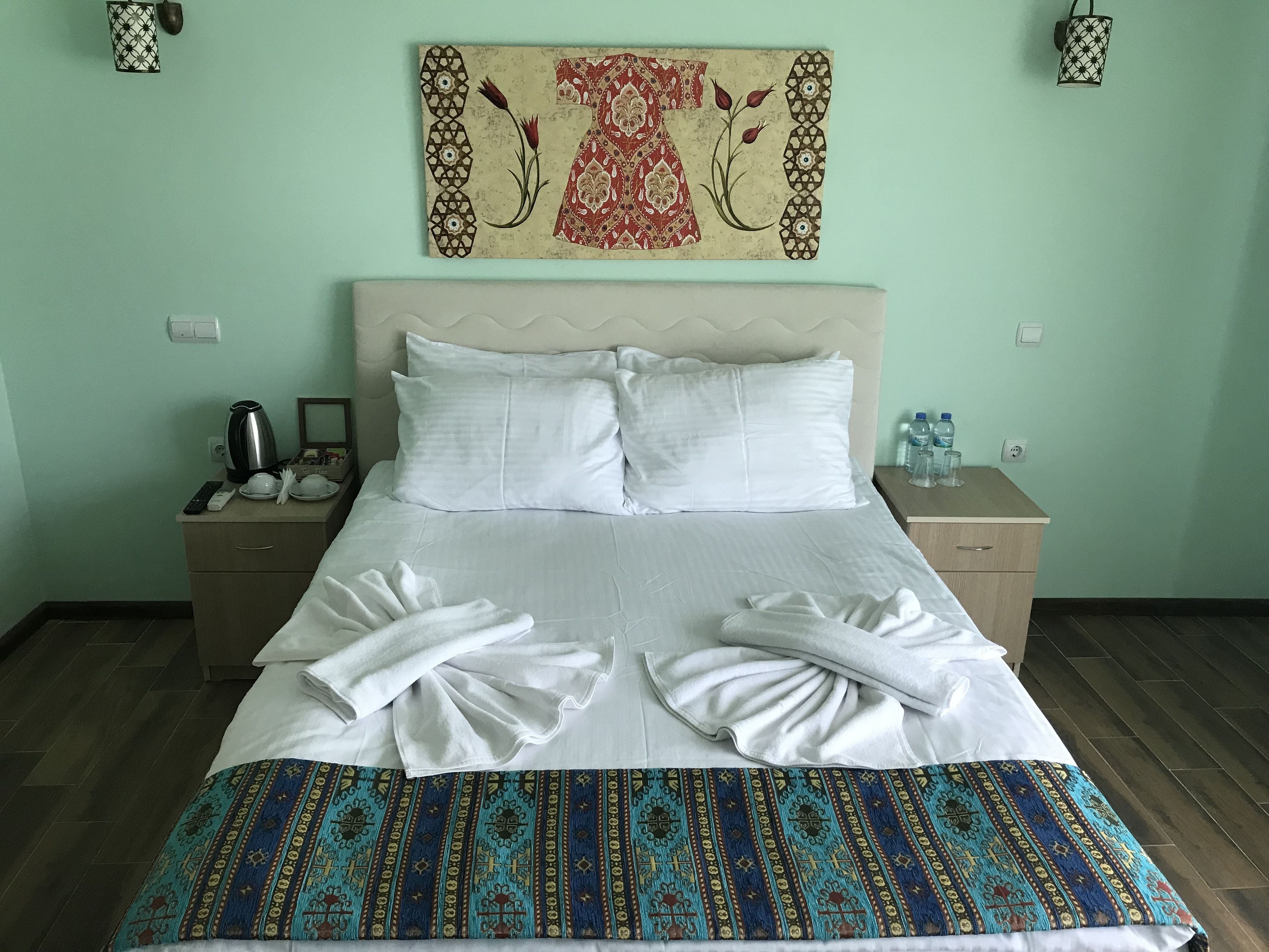 Een netjes opgemaakt bed met witte lakens en een bedekte deken, een nachtkastje met een waterkoker en een afstandsbediening, en een kunstwerk aan de muur boven het bed.