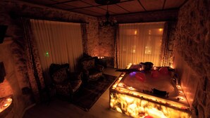 A dimly lit room with a stone wall, a fireplace, a large bathtub with candles, and two ornate chairs. - Kemal Stone House Hotel (Nevsehir)