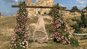 A rustic outdoor setting with a stone structure in the background, a wooden swing, and pink flowering plants. - Kemal Stone House Hotel (Nevsehir)