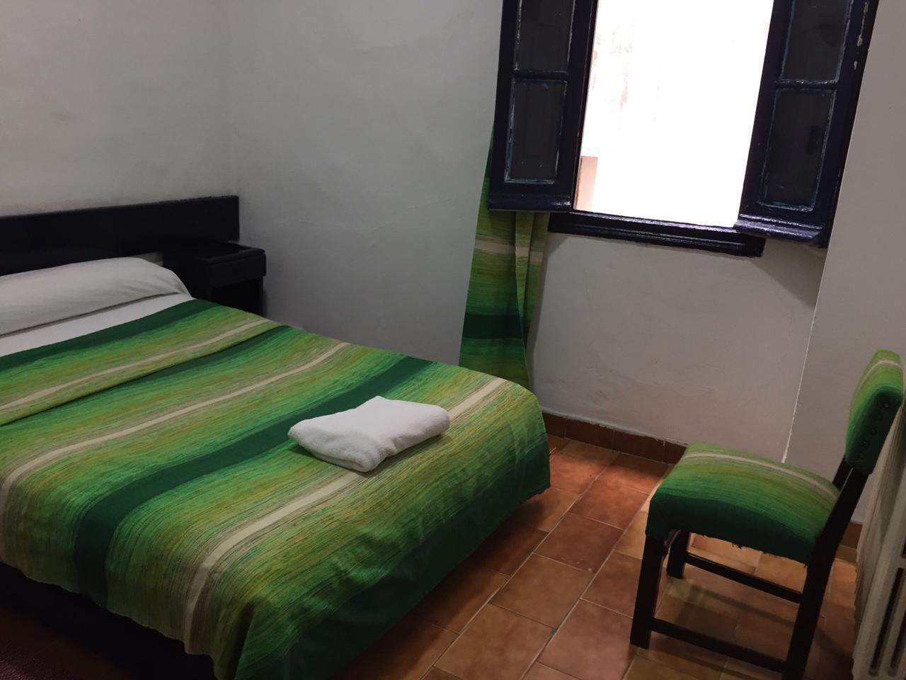 A small, clean room with a bed, a chair, and a window with shutters.