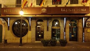 En hotellfasad med en skylt som visar texten ”HOTEL RECREA” och arabisk text. Framför finns flaggor, ett stort runt fönster och krukväxter.