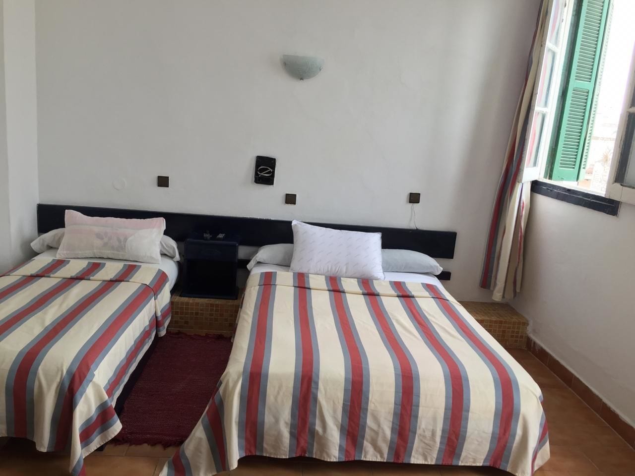 A room with two beds, each with striped bedding, a small rug, a nightstand, and a wall-mounted light fixture.