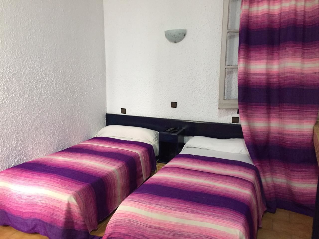 Ein Zimmer mit zwei Einzelbetten, einem Nachttisch, einer Wandleuchte und einem Fenster mit Vorhang.