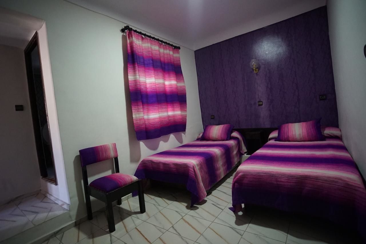 Quarto com duas camas de solteiro, uma cadeira e janela com cortina roxa.