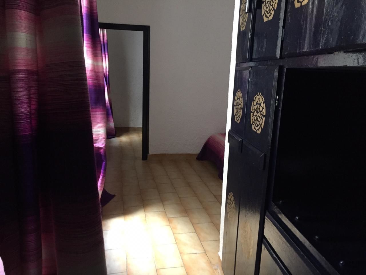 A room with a tiled floor, a bed with a purple blanket, and a black wardrobe with decorative patterns.