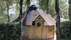Een klein houten huisje met een zeszijdige deur en een schuin dak, omringd door weelderige begroeiing en bomen.