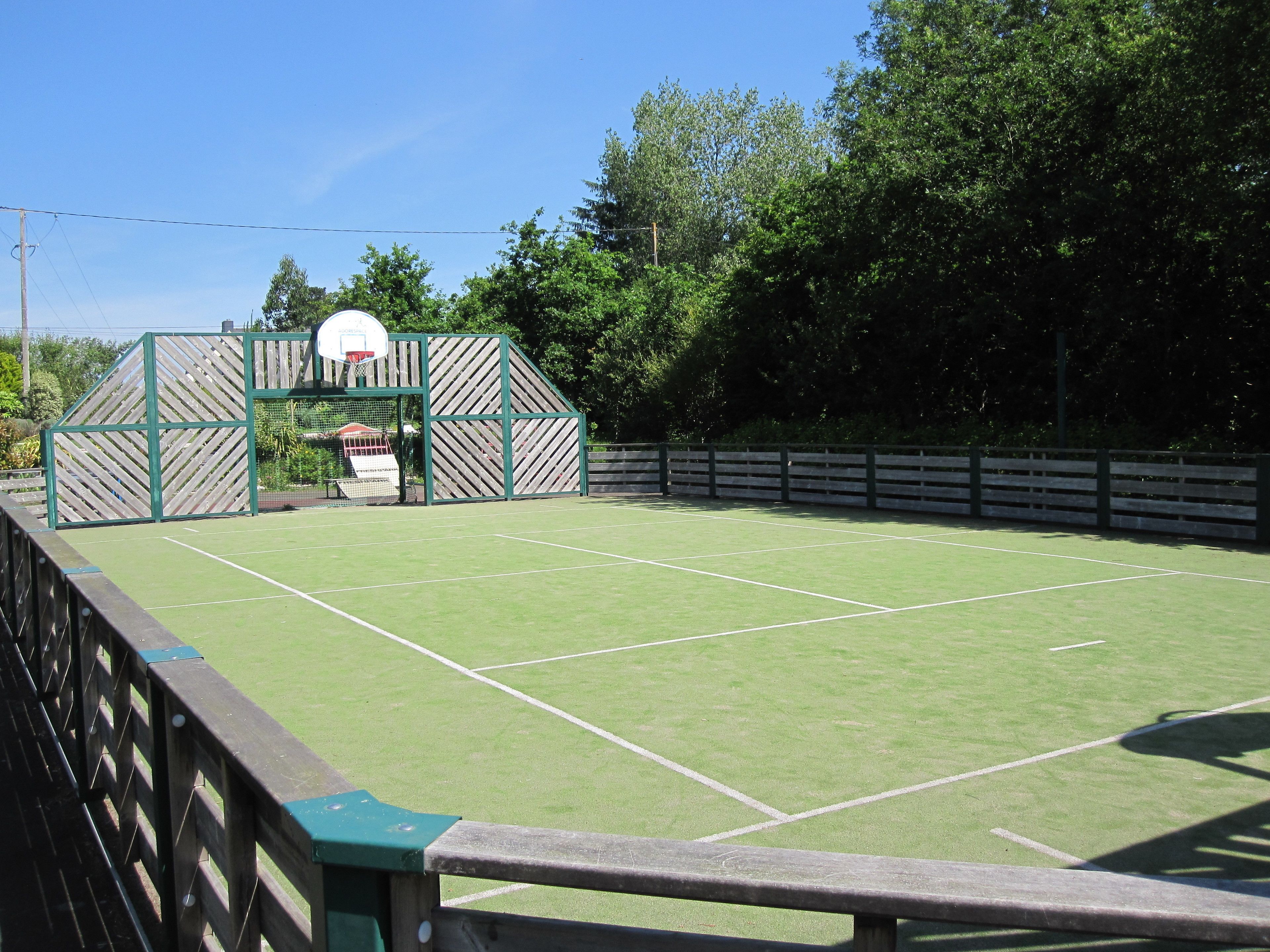 Un court de tennis en plein air, entouré d’une clôture en bois, avec un panier de basket.