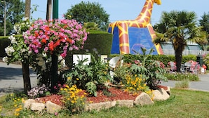 Een tuin met een grote girafssculptuur, diverse bloeiende planten en een strak bijgesnoeid haag.