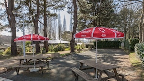 Een picknickplaats met houten tafels en banken, rode parasols met logo's, en een grindpad.