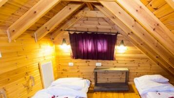 A small, cozy attic bedroom with wooden walls and a sloped ceiling. There are two beds with white linens and a small table in the center.