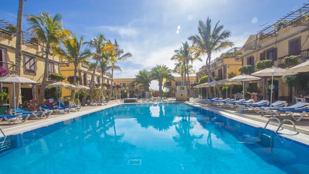 Une piscine entourée de palmiers et d’immeubles, avec des chaises longues et des parasols.