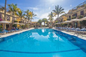 Une piscine entourée de palmiers et d’immeubles, avec des chaises longues et des parasols.
