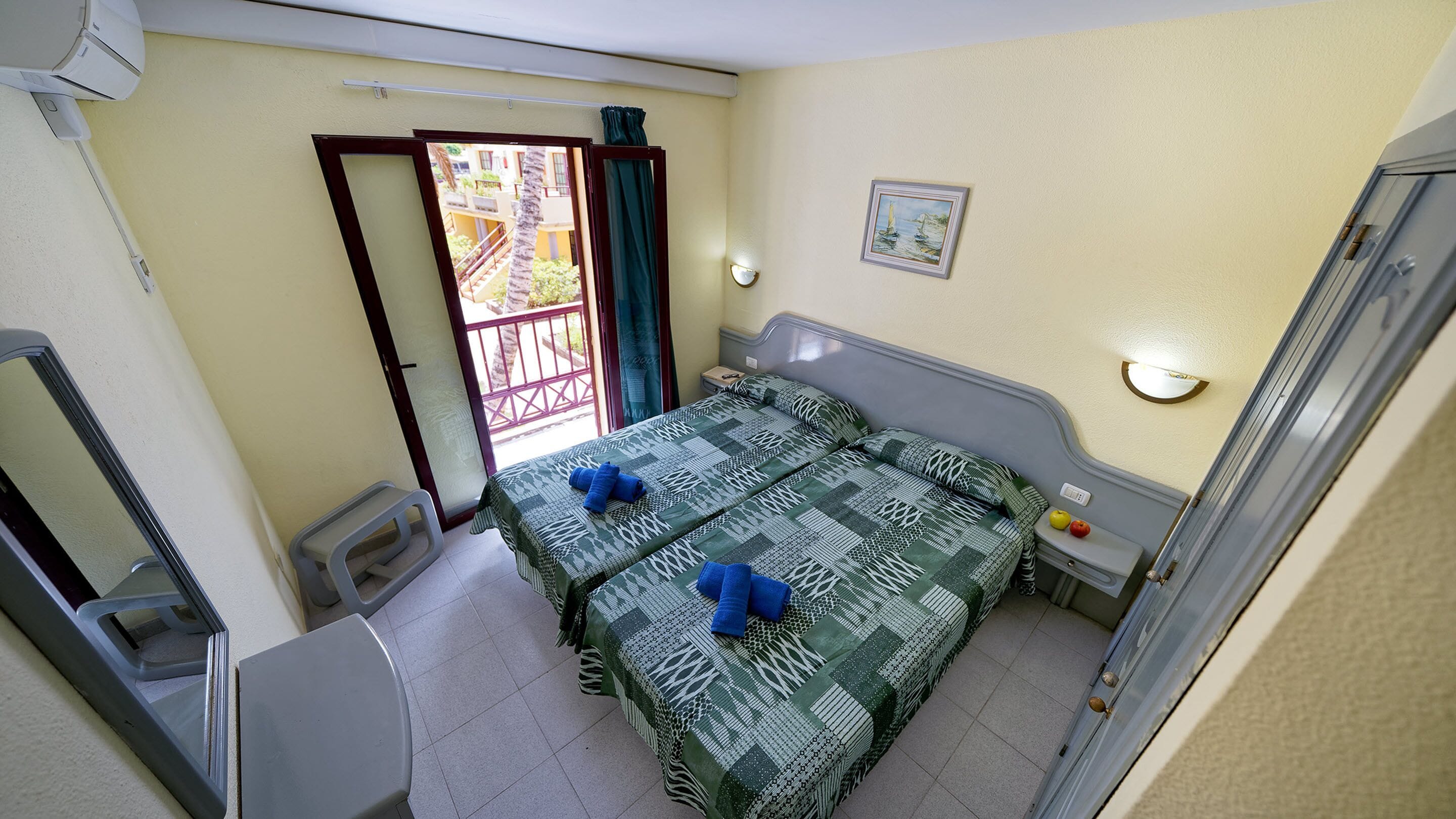 A room with a bed, a small table, a chair, a window with a view of a balcony, and a wall-mounted light fixture.