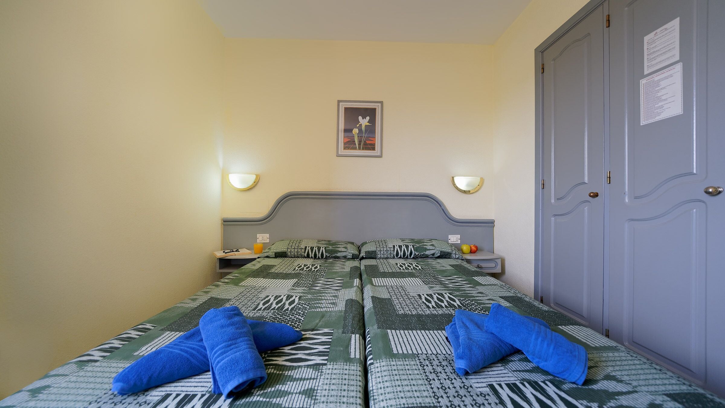 A single bed with patterned bedding, two bedside tables with lamps, a framed picture on the wall, and a wardrobe with a door.