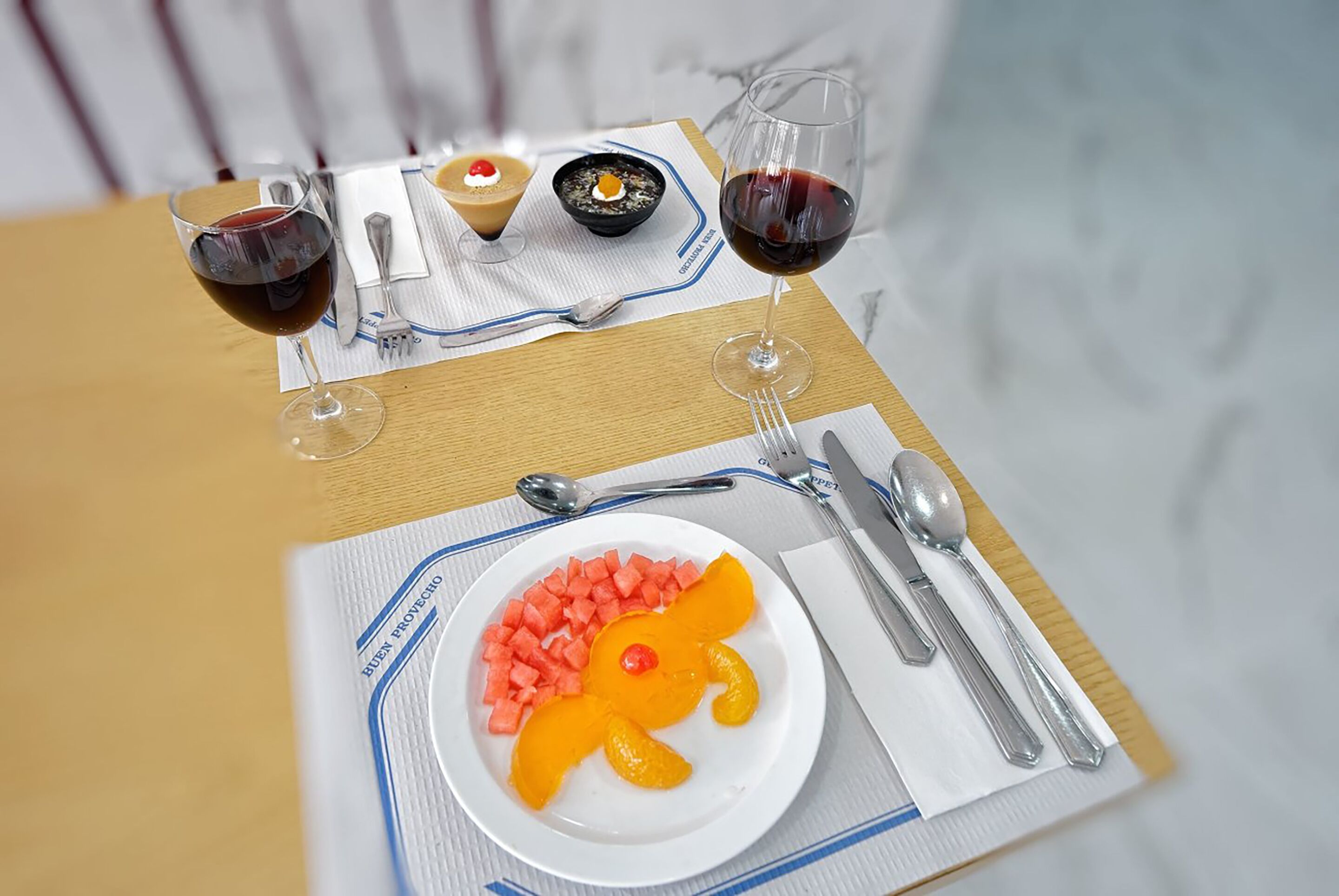 A dining table with a dessert plate, two glasses of red wine, and a bowl of dessert on a placemat.