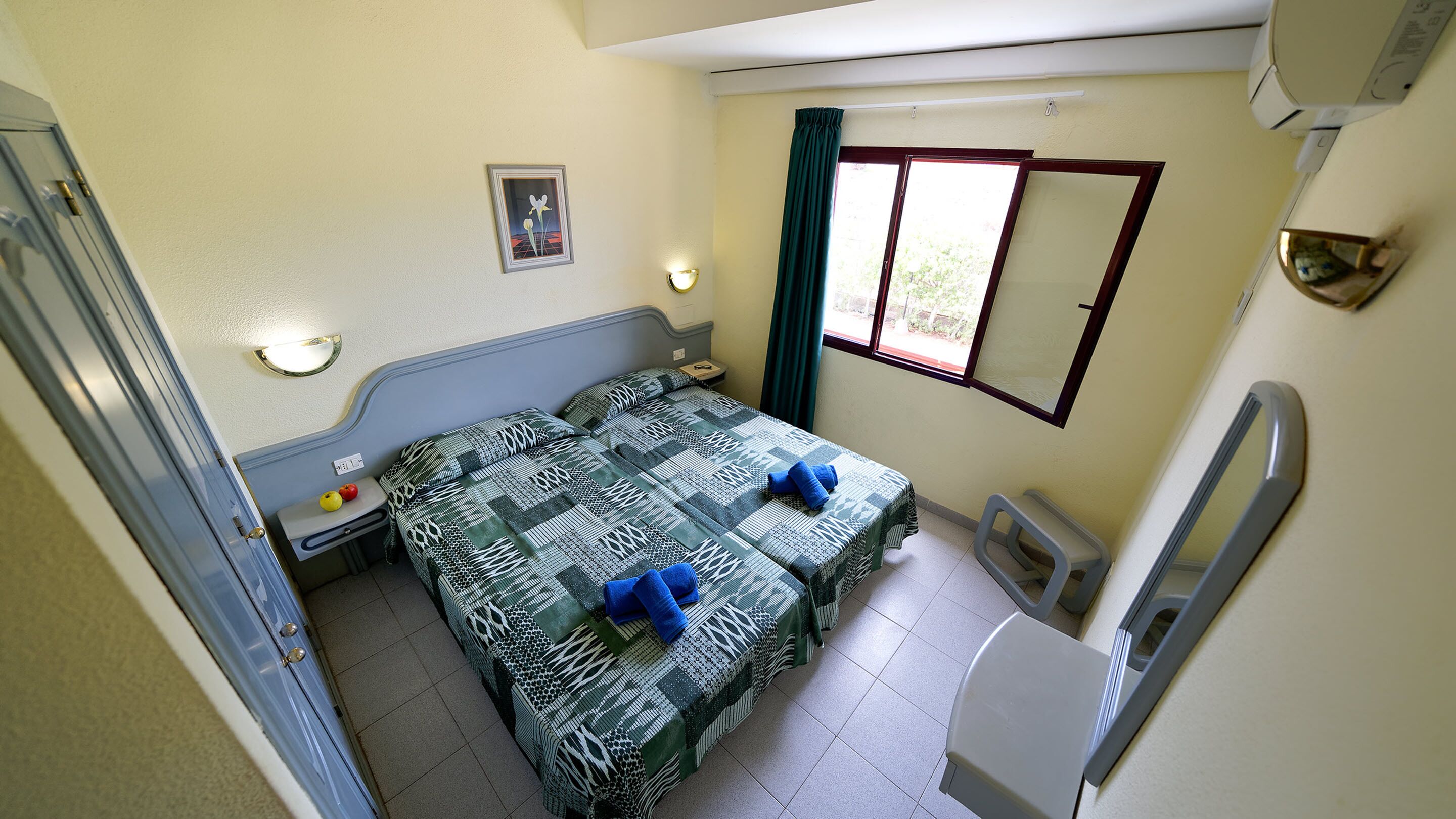 A compact room with two beds, a small table, a wall-mounted light, a window with curtains, and a mirror.