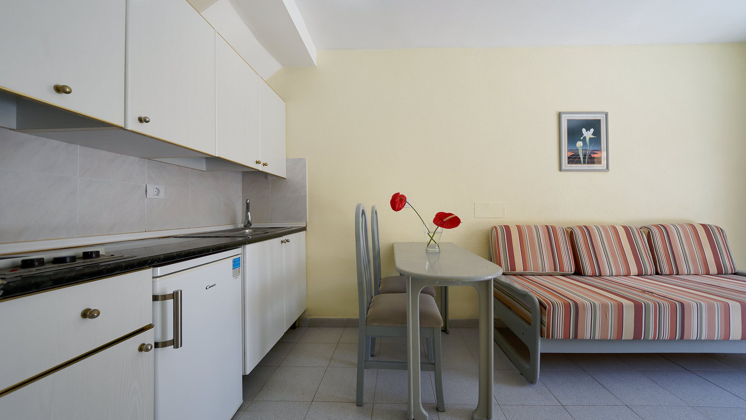 Une cuisine compacte avec des meubles blancs et un plan de travail noir, une petite table à manger avec deux chaises et un canapé agrémenté de coussins à rayures.