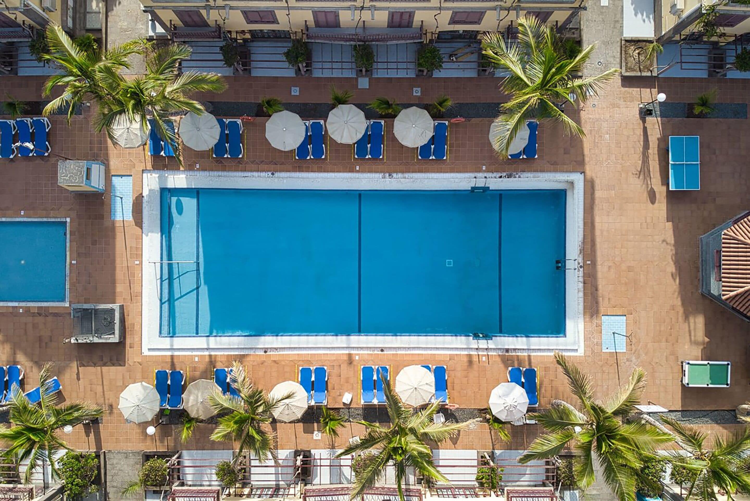 Een zwembad op het dak met blauw water, omringd door ligbedden, parasols en palmbomen.