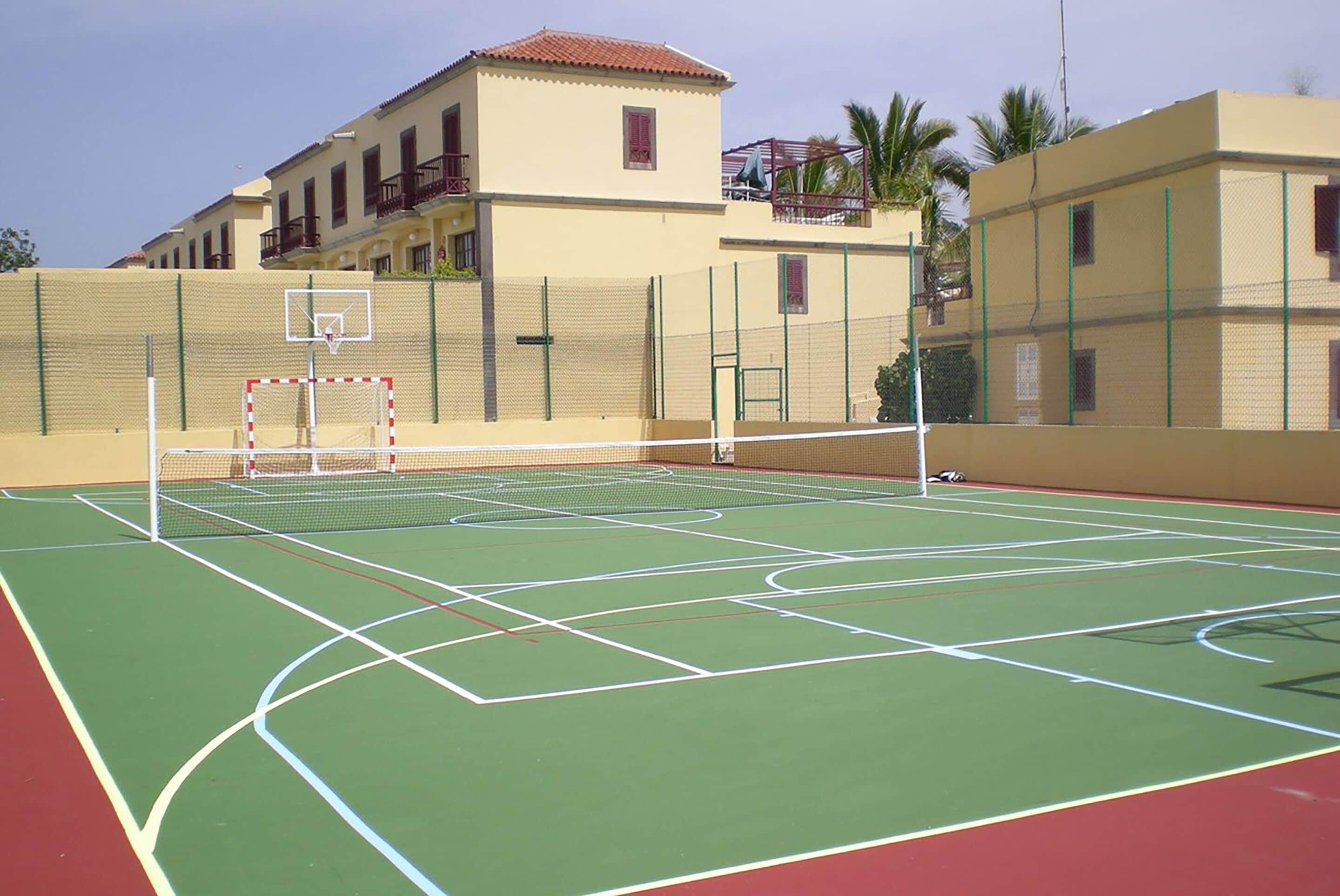 Un terrain de tennis extérieur, doté d’un filet, entouré d’une clôture et avec des bâtiments en arrière-plan.