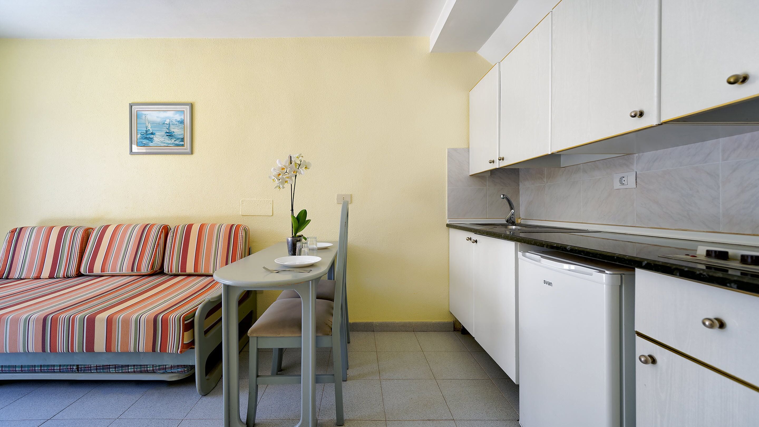 Una kitchenette compacta con armarios blancos, una pequeña mesa de comedor y un sofá con cojines a rayas.