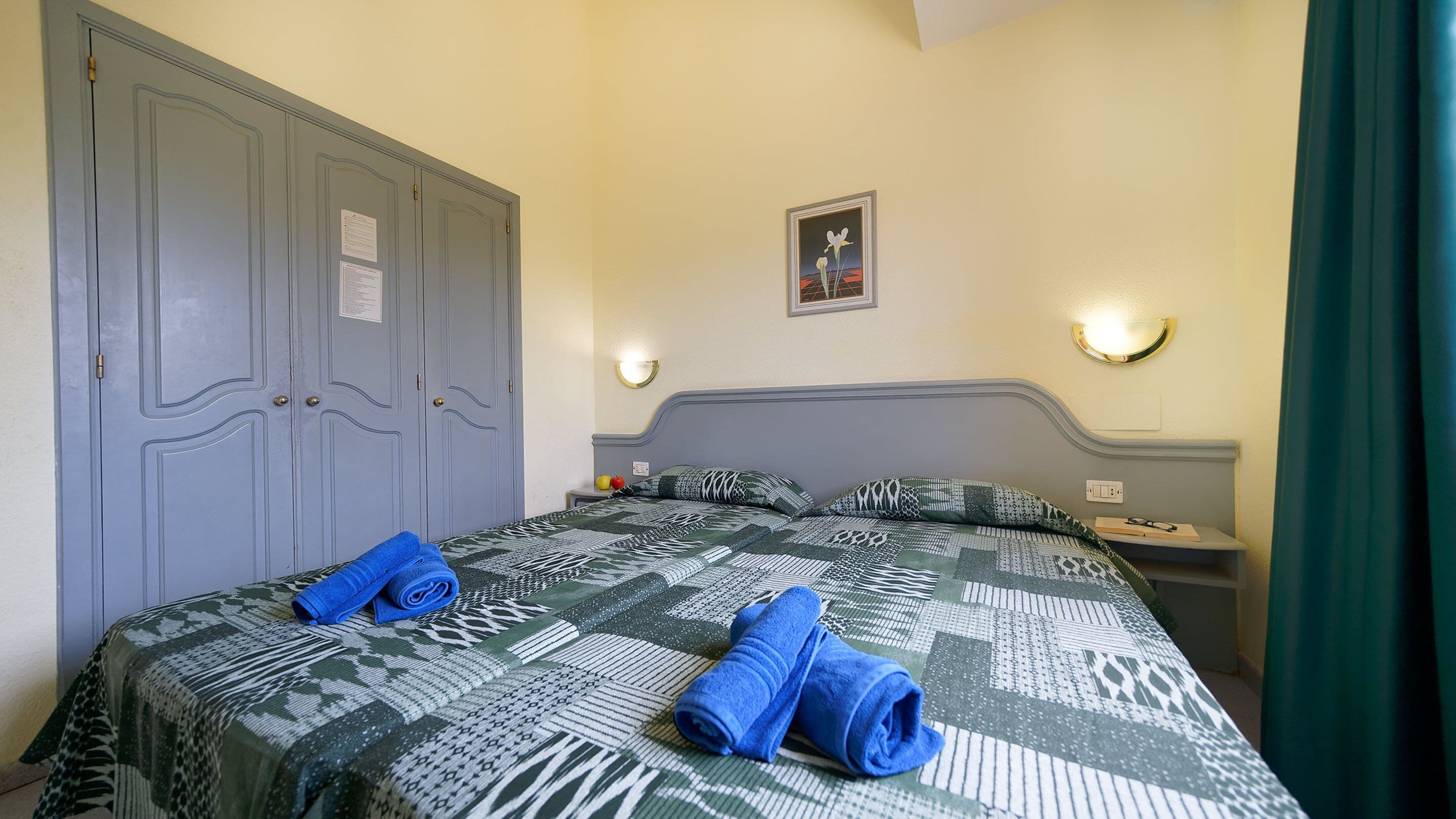 A bedroom with a large bed, two pillows, a blue towel, a nightstand, a wall-mounted light, and a door with two locks.