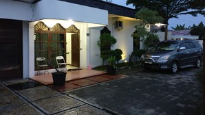 A house with a paved driveway, a car parked in the driveway, and a small garden area with a chair and potted plants. - Wisma Liontine (Palangkaraya)
