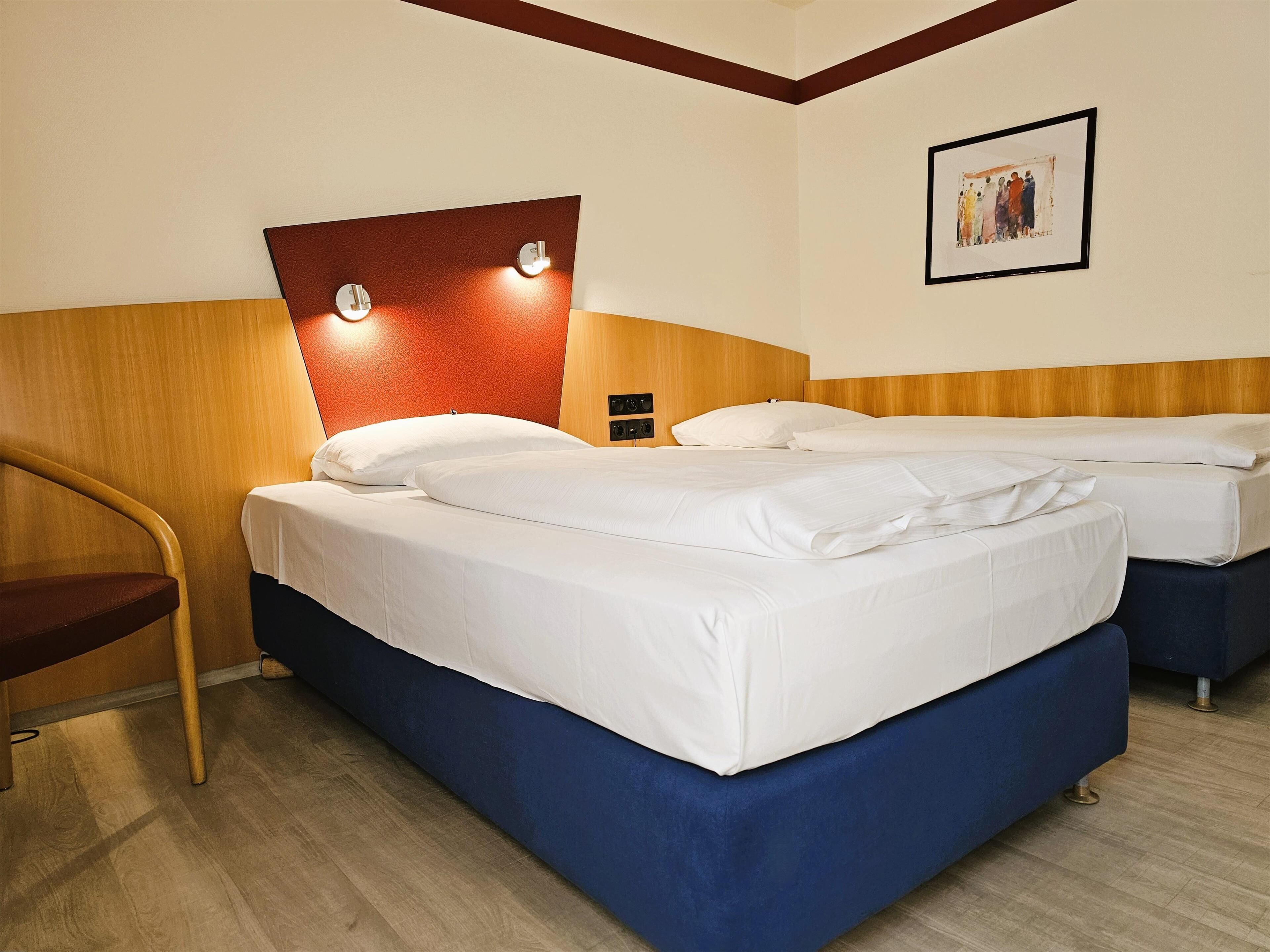 Habitación de hotel con cabecera de madera, dos camas con sábanas blancas, una silla de madera y un cuadro en la pared.