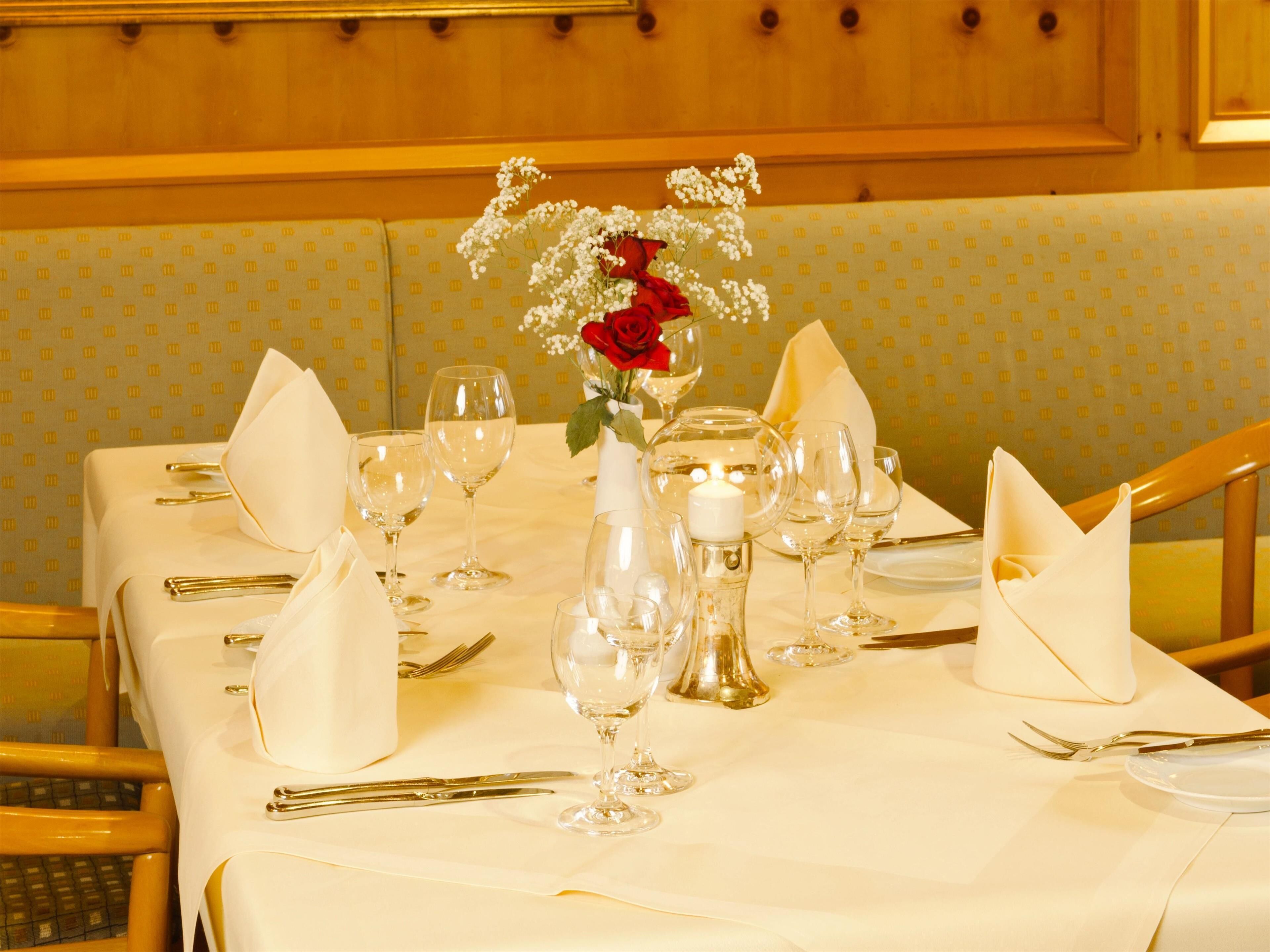 Una mesa de comedor dispuesta con servilletas blancas, copas de vino y cubiertos, con un jarrón de flores y una vela como centro.