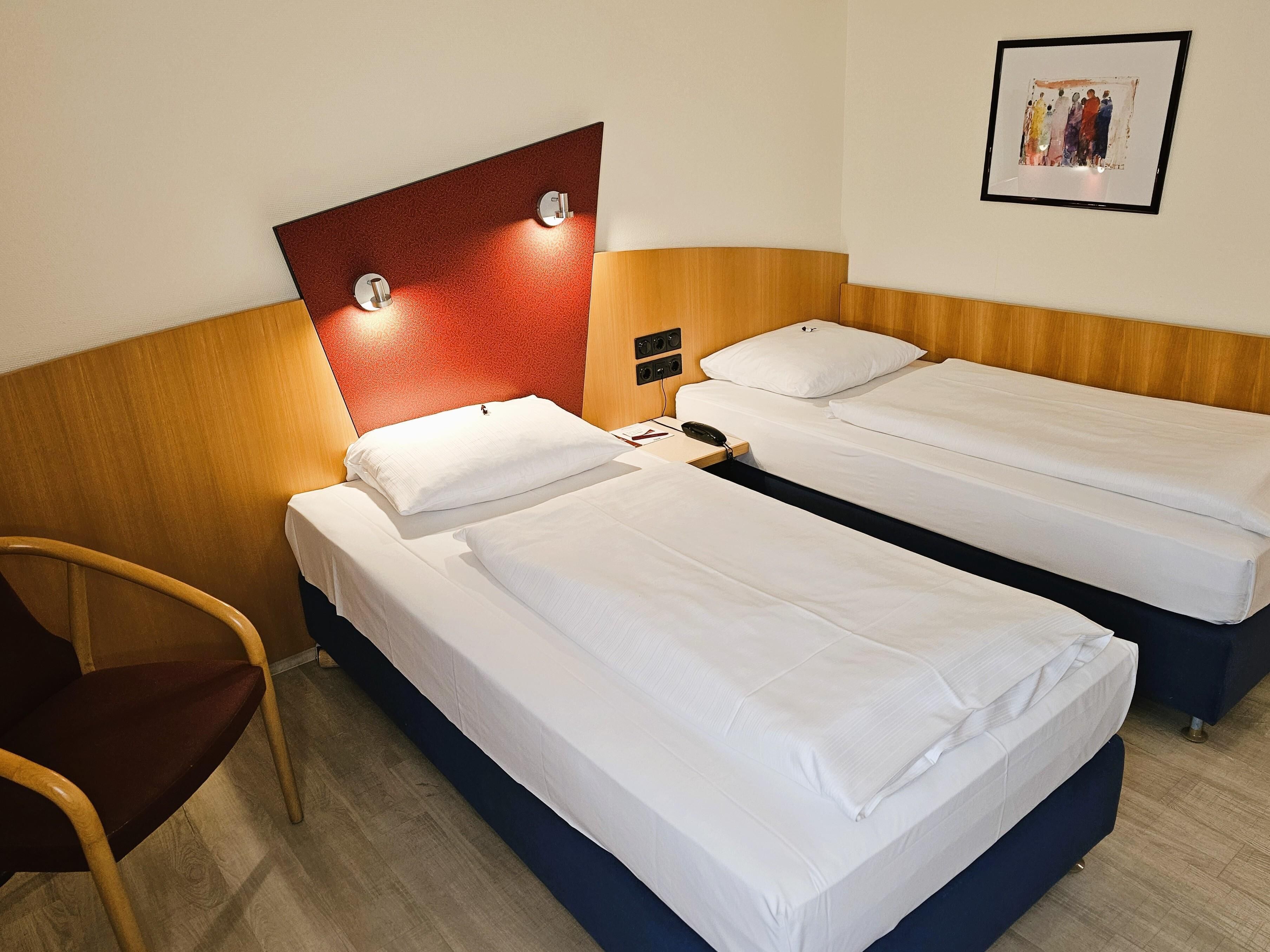A hotel room with two single beds, a wooden headboard, a wooden chair, and a framed picture on the wall.