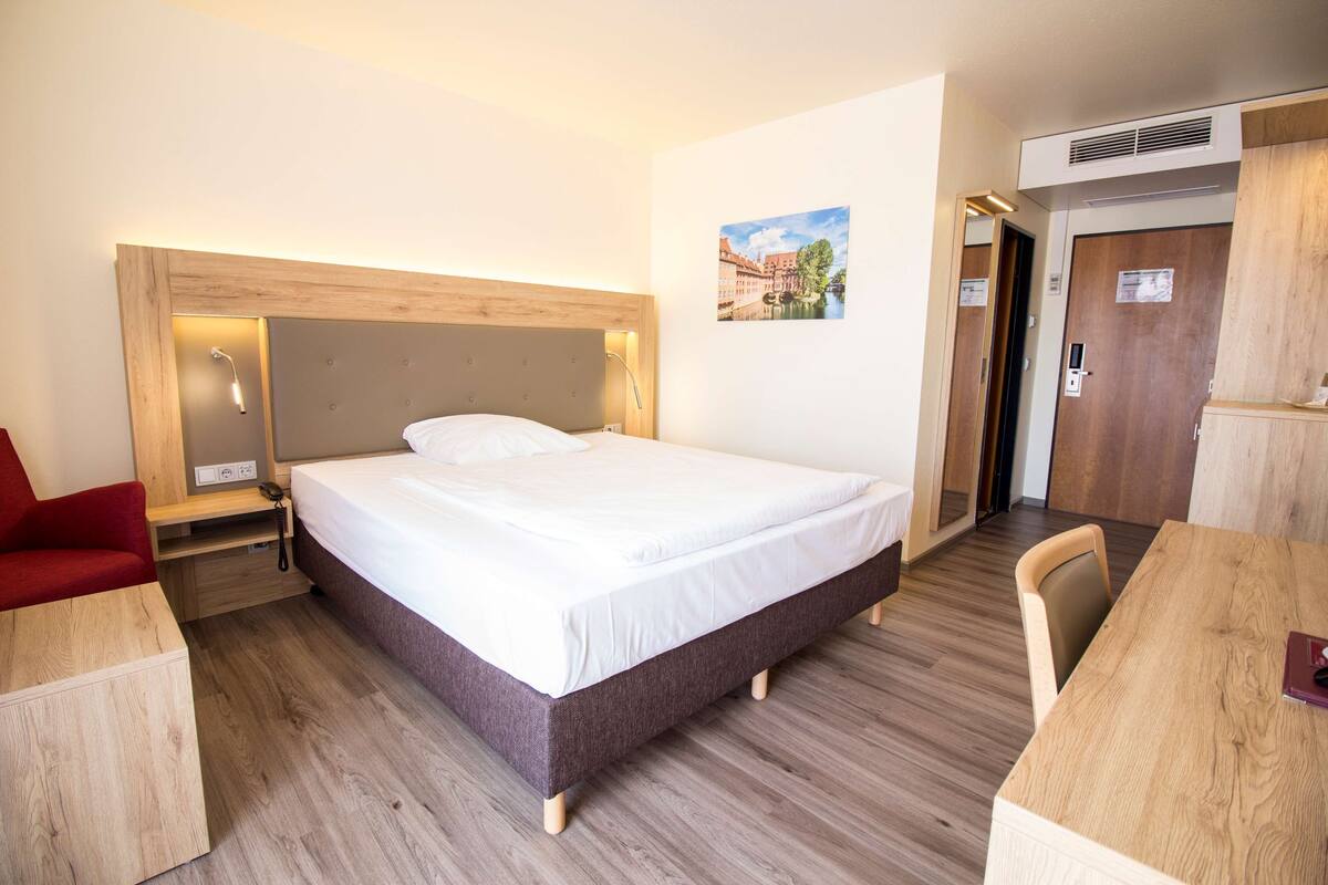 Una habitación de hotel con una cama grande, cabecera de madera, escritorio, silla y un pequeño sofá rojo.