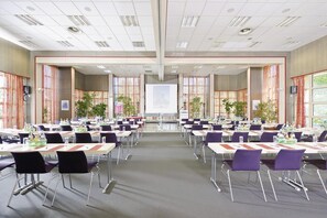Ein Konferenzraum mit mehreren Tischen und Stühlen, großen Fenstern mit Vorhängen und einer Projektionsleinwand an der Vorderseite.