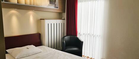 Habitación de hotel con cama, una silla, un radiador y un ventanal con cortinas.
