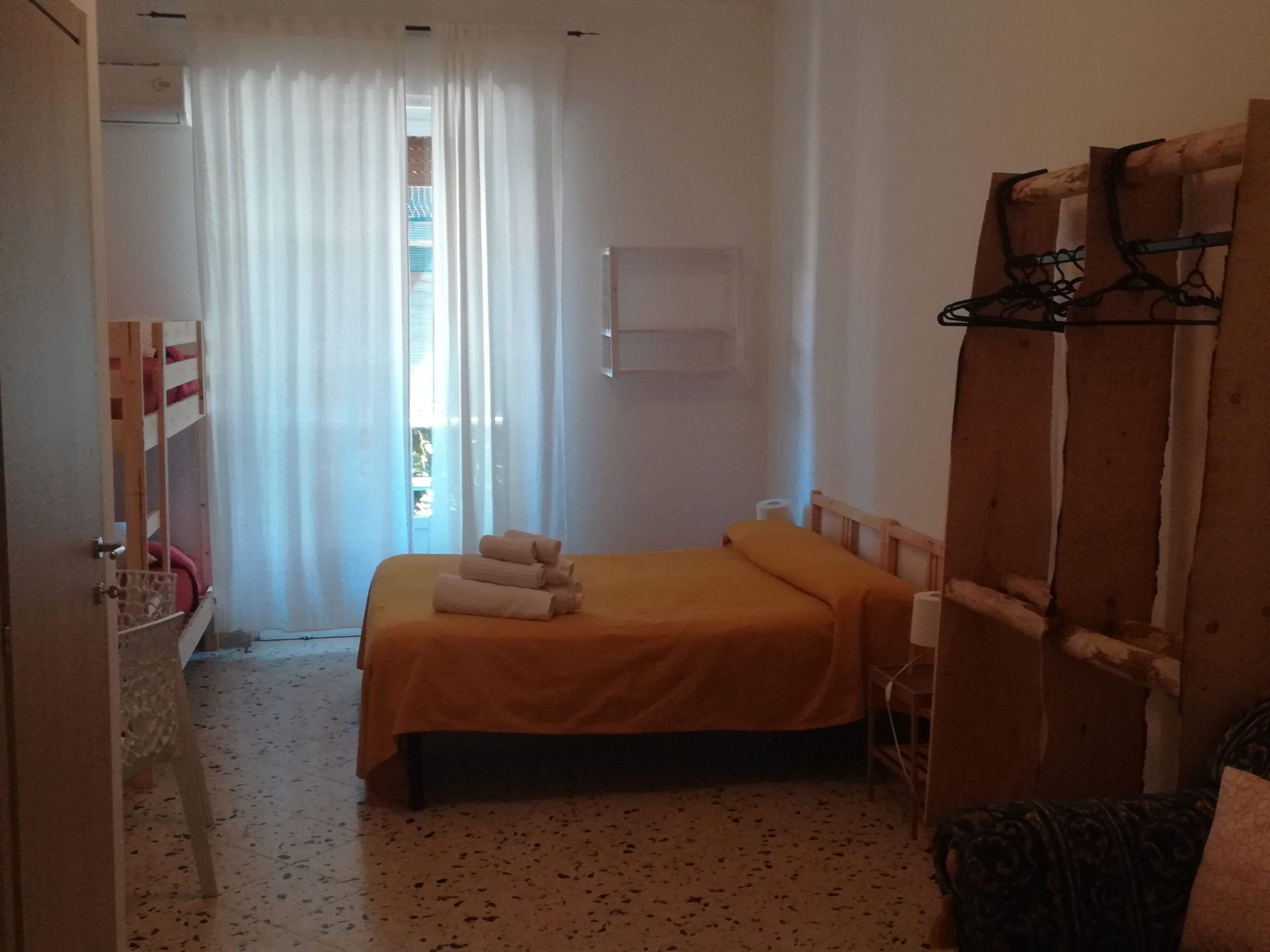 Quarto pequeno e simples com uma cama, um guarda-roupa de madeira, uma janela com cortinas e uma porta.