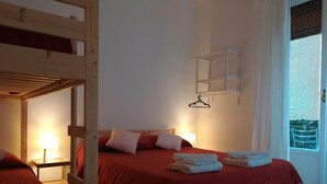 Une chambre avec un lit superposé, un lit simple avec une literie rouge, une table de chevet avec une lampe et une fenêtre avec des stores.