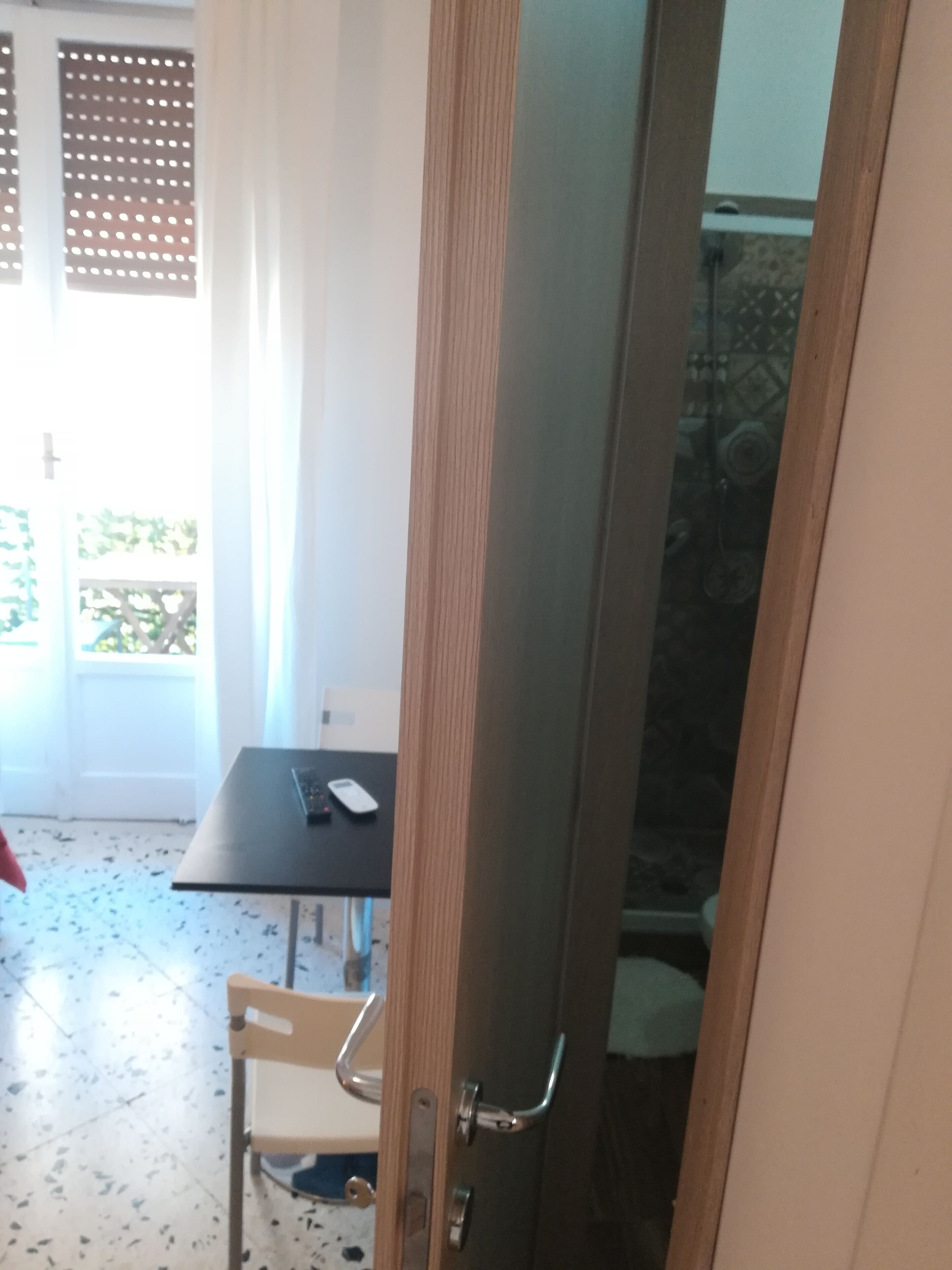 A room with a wooden door, a small table with a chair, and a bathroom visible through a glass door.