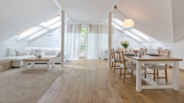 Un loft spacieux avec un plafond haut, un plancher en bois, un coin repas avec une table et des chaises en bois, un ensemble de canapés et une vue dégagée sur l’extérieur grâce à une grande fenêtre.