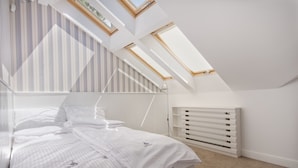 Une chambre avec un plafond en pente, des fenêtres de toit, un lit avec une literie blanche, un radiateur et un papier peint à rayures.
