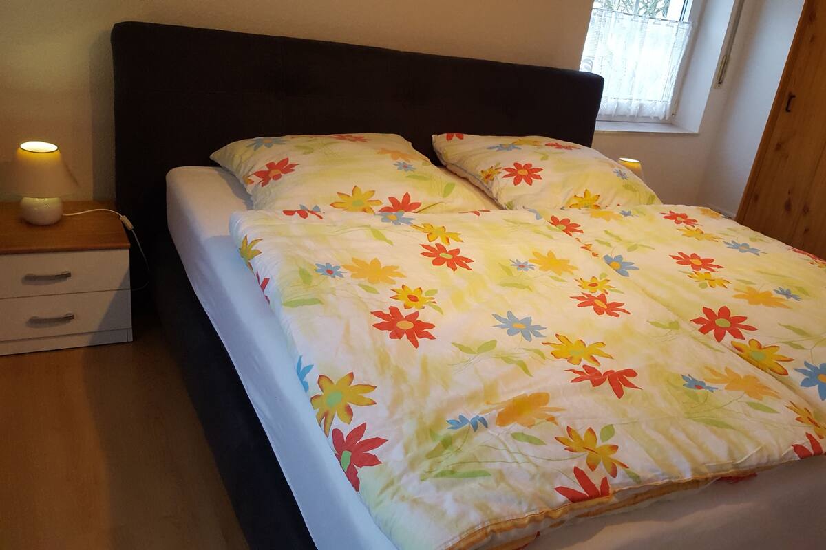Una cama con colcha estampada, cabecero de madera, mesitas de noche con lámparas y una ventana con cortinas.