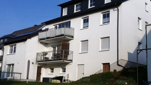Un edificio residencial de varios pisos con un balcón en el segundo nivel, una puerta de garaje y un pequeño jardín.