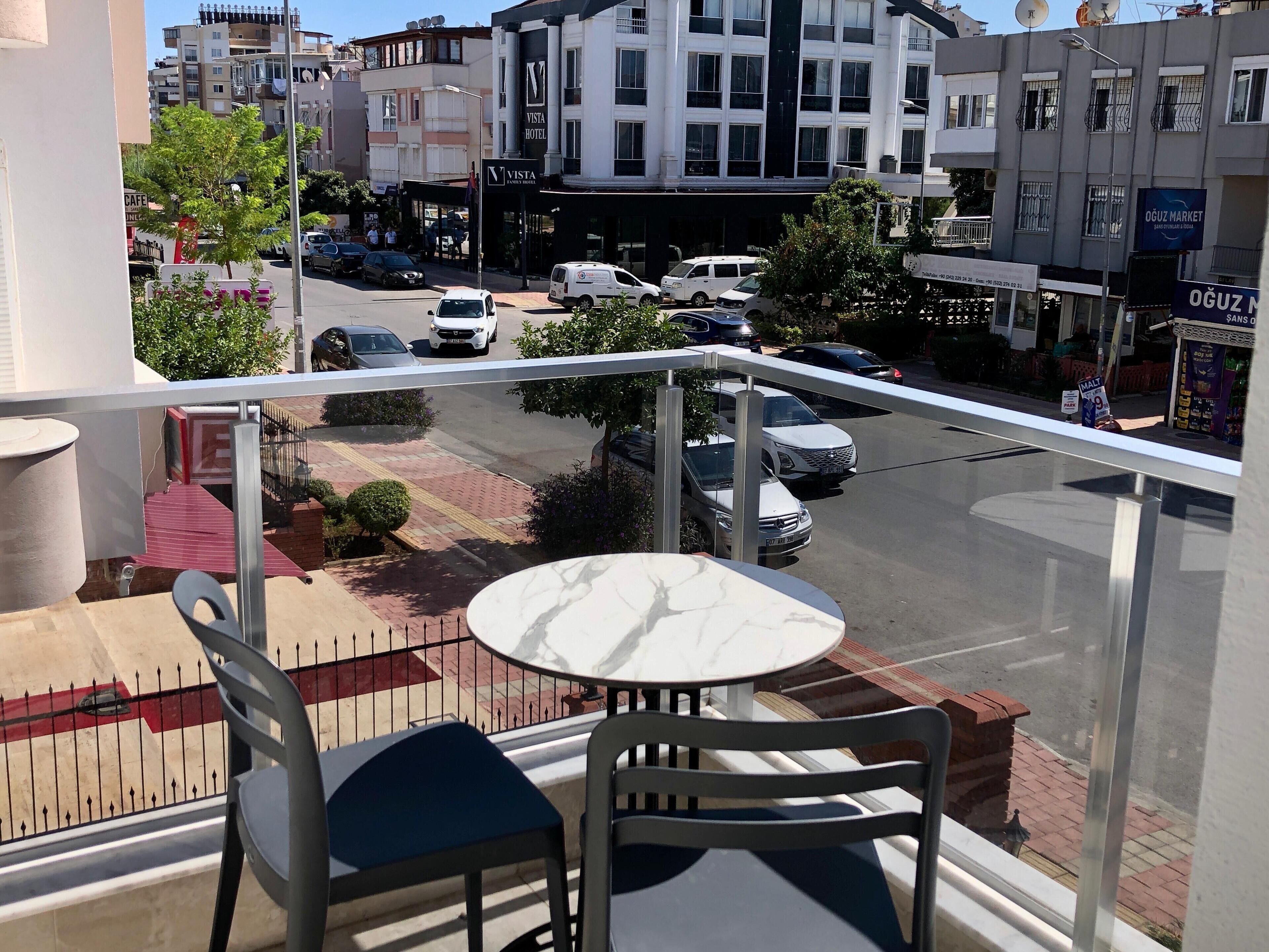 Un balcón con mesa y sillas, con vistas a una calle con coches aparcados y edificios, incluyendo un hotel y un mercado.