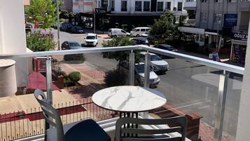 Un balcón con mesa y sillas, con vistas a una calle con coches aparcados y edificios, incluyendo un hotel y un mercado.