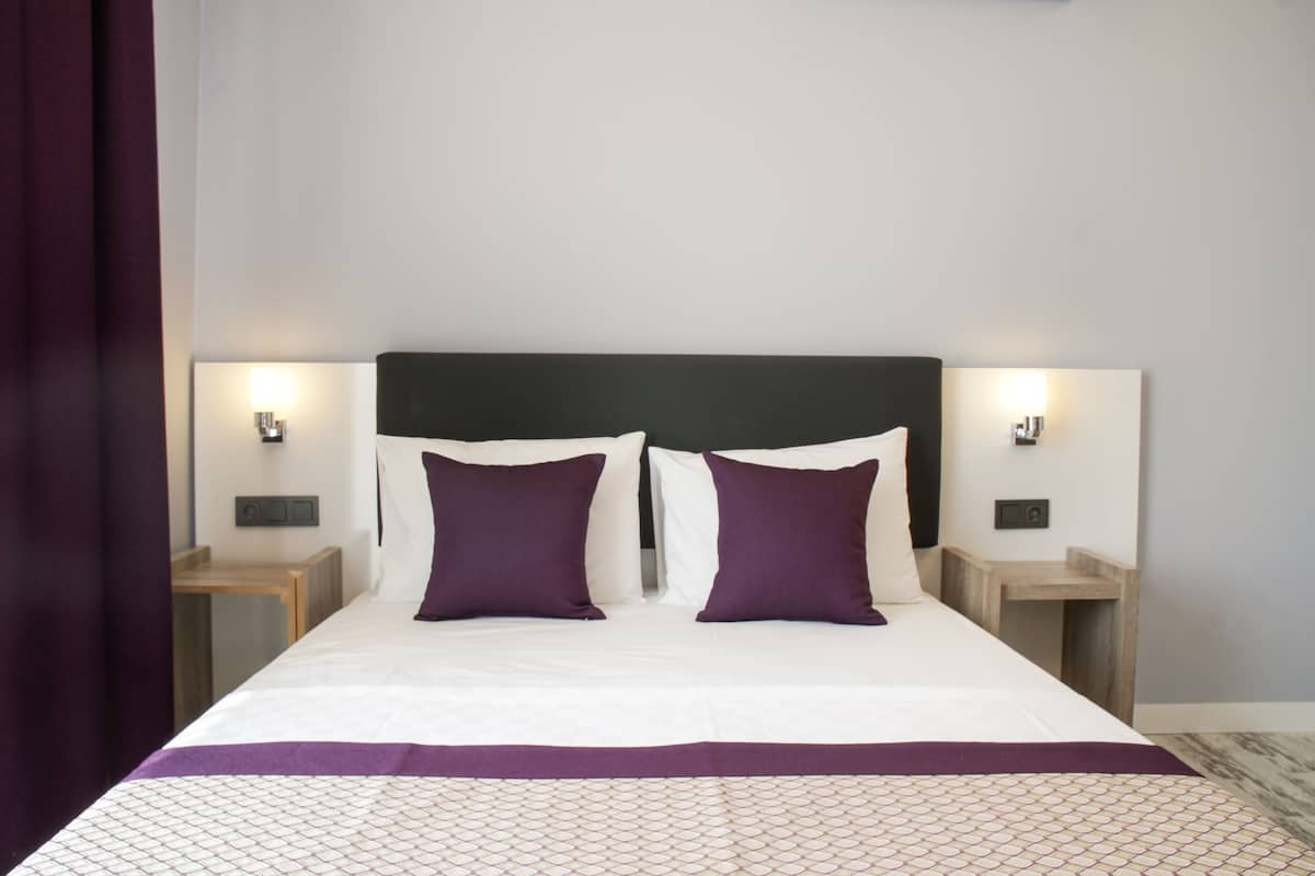 A neatly made bed with a white and purple bedspread, two matching pillows, and a dark headboard. There are two bedside tables with lamps on either side of the bed.