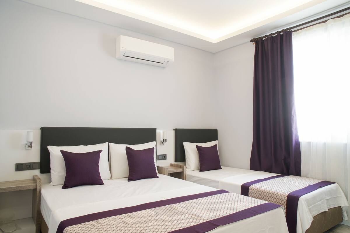 A hotel room with two beds, a ceiling air conditioner, a nightstand, and a window with curtains.