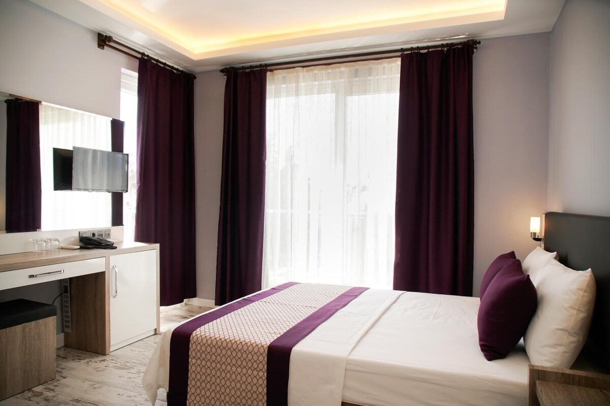 A hotel room with a bed, a desk with a computer, a TV, and a window with curtains.