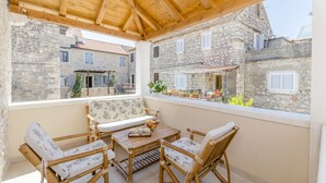 A covered outdoor seating area with wicker furniture and a table, overlooking a courtyard with stone buildings and a balcony with potted plants. - House Morko1 (Vrboska)