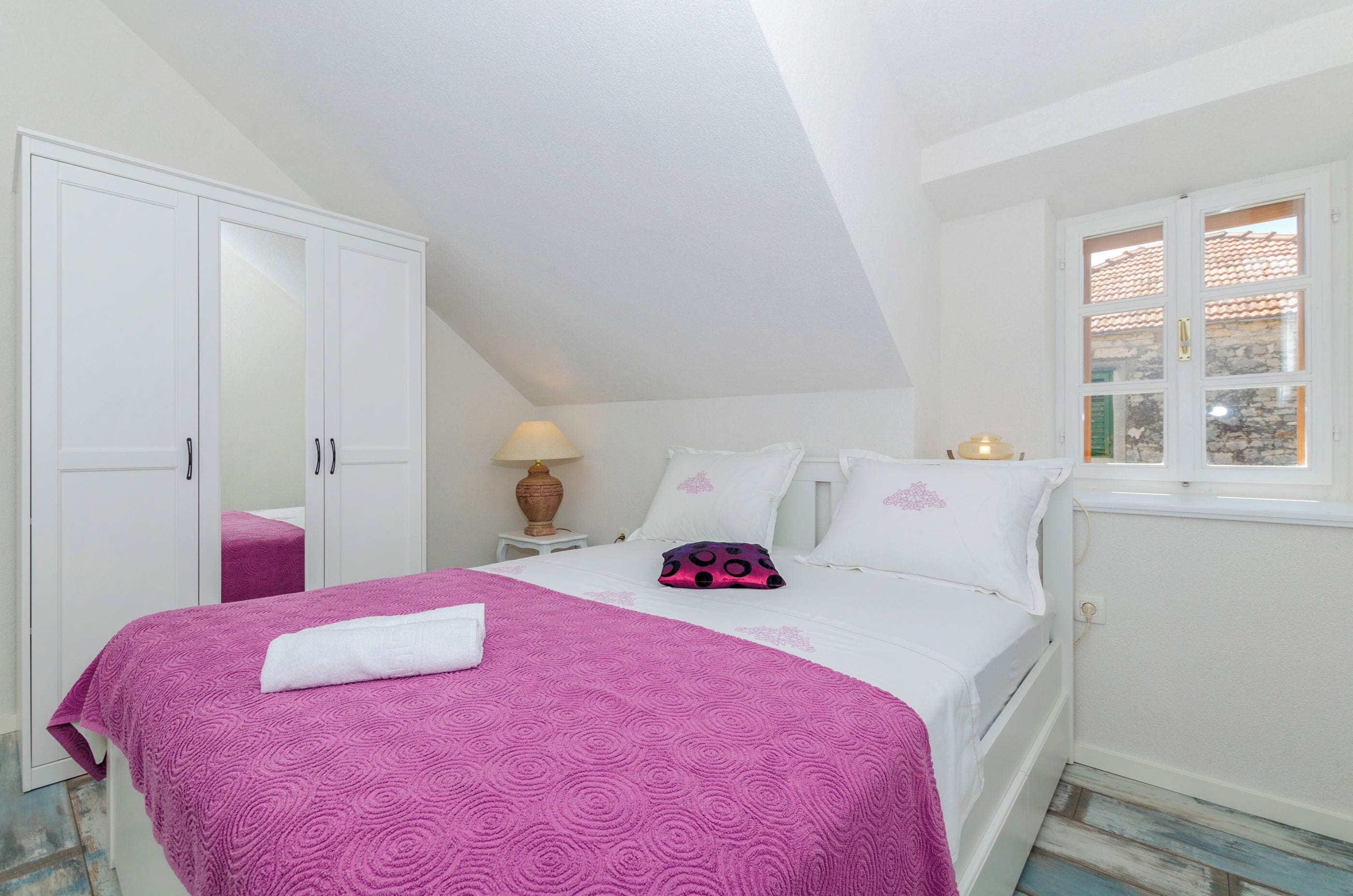 Ein Schlafzimmer mit Bett, Kleiderschrank, Nachttisch mit Lampe und Fenster mit Blick nach draußen.