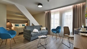 A modern hotel room with a grey sofa, blue armchairs, a wooden floor, a bed with a headboard, and a large window with curtains. - EA Hotel New Town (Prague)
